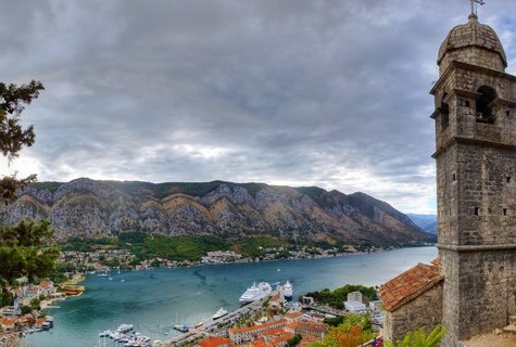 Panoramic view of Kotor Bay showing a stone bell tower on the right, cruise ships docked in the harbor, red-roofed buildings of the old town, turquoise water, and steep mountains under cloudy skies