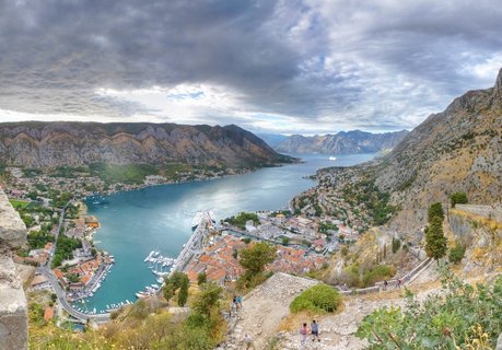 Panoramic view of Kotor Bay in Montenegro showing the old town with red roofs, cruise ships in the harbor, surrounding mountains, and stone fortress walls with hiking paths in the foreground under a cloudy sky