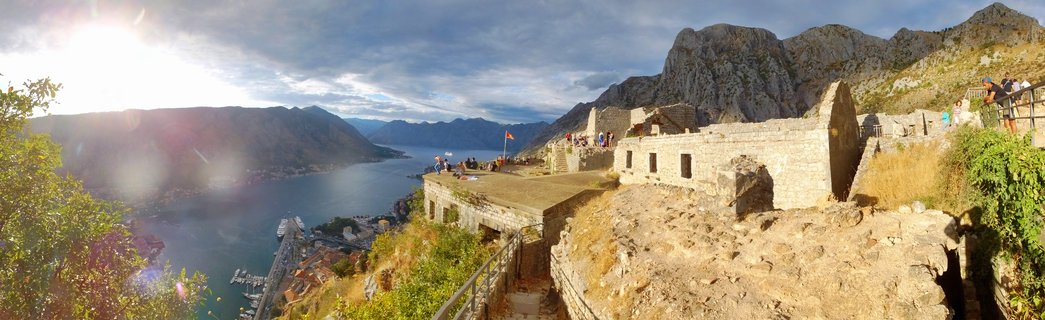 Panoramic view from medieval stone fortress walls showing tourists, the Bay of Kotor, red-roofed old town, and mountains under a partly cloudy sky with bright sun