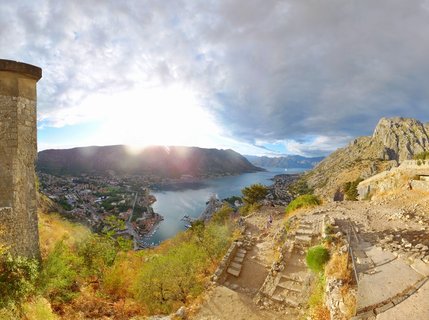 Panoramic view from Kotor Fortress showing stone hiking path, medieval walls, Bay of Kotor with cruise ship, Old Town with red roofs, and surrounding mountains under cloudy sky with sun breaking through