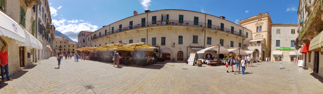 Wide panoramic view of a historic European town square with stone buildings, outdoor restaurant seating under yellow umbrellas, people walking on cobblestone pavement, blue sky with scattered clouds
