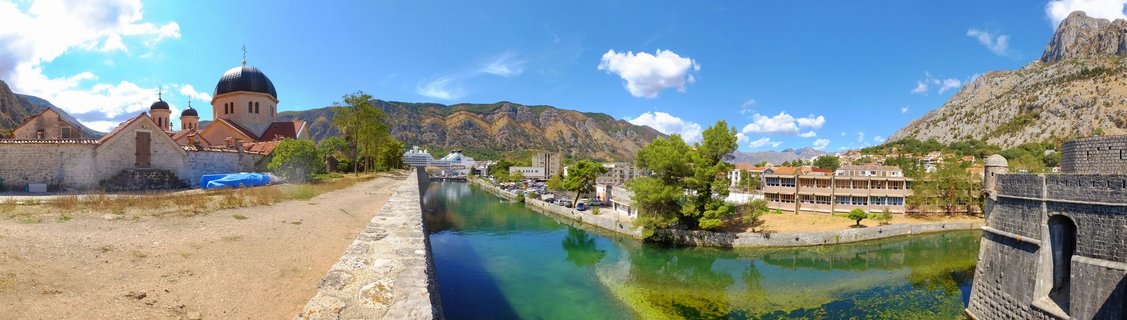 Panoramic view of a historic canal in Budva, Montenegro, showing an Orthodox church with domes on the left, turquoise water flowing through stone embankments, medieval fortress walls on the right, and mountains in the background under blue sky with scattered clouds