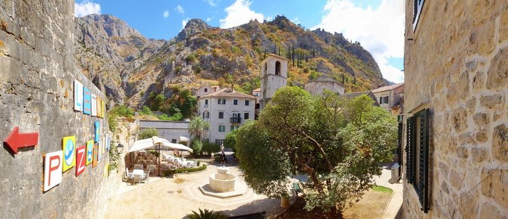 Stone plaza in historic old town with church bell tower, outdoor café seating, central fountain, and mountains in background under blue sky