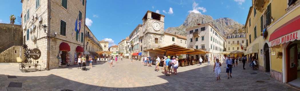 Panoramic view of a historic stone town square with a clock tower, surrounded by Mediterranean buildings, tourists walking, outdoor cafes, and mountains in the background under blue sky