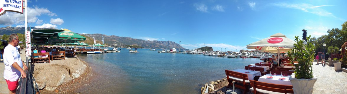 Panoramic harbor scene showing waterfront restaurant terraces with tables and umbrellas on both sides of a marina filled with boats, mountains visible in the distance under blue sky
