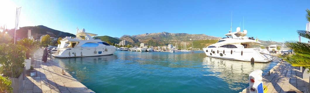 Panoramic view of a marina in Budva, Montenegro showing two large white luxury yachts docked at a stone pier with turquoise water, palm trees, and mountains in the background under a clear blue sky