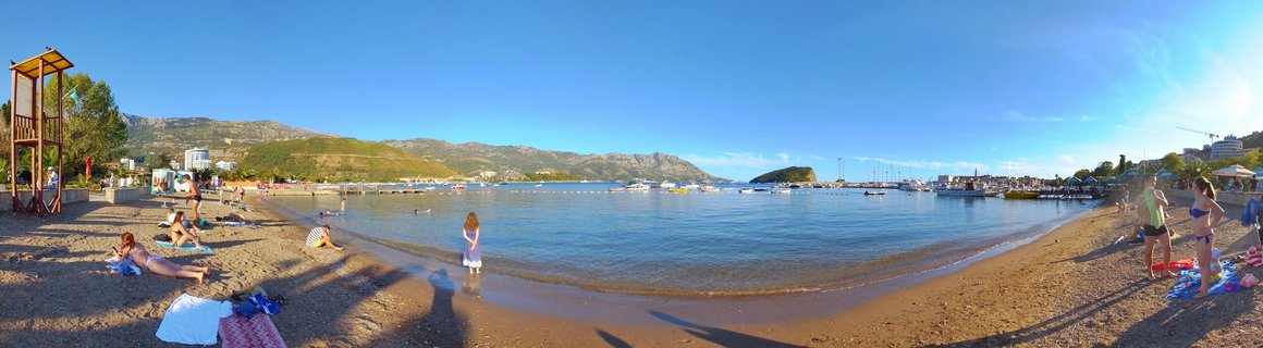 Panoramic beach scene showing people sunbathing and swimming at Budva beach in Montenegro, with a wooden lifeguard tower on the left, boats in the harbor, and mountains in the background under a clear blue sky
