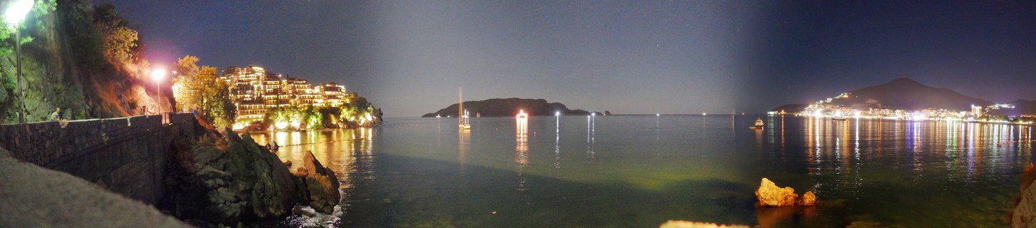 Panoramic night photograph of Budva, Montenegro showing illuminated coastal buildings on hillsides, calm Adriatic Sea with boat light reflections, and distant island under dark sky