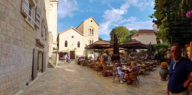 Wide street view of a historic stone plaza in Old Town Budva with people seated at outdoor café tables under brown umbrellas, a cream-colored church with arched windows in the center background, stone buildings with shutters on the left, and blue sky with scattered clouds above.