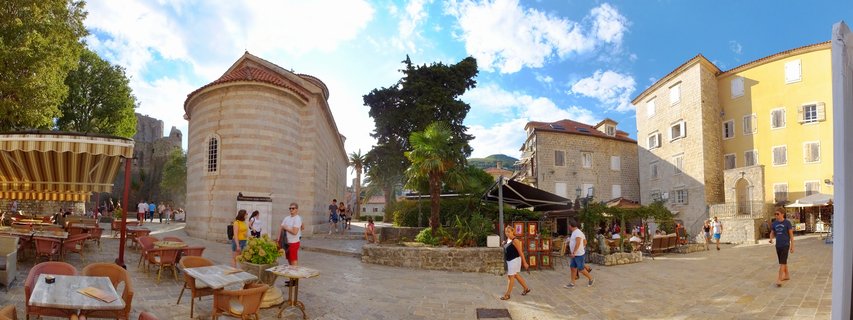 Wide panoramic view of a stone-paved town square in Budva, Montenegro, showing a round stone church with red-tiled roof, yellow and stone buildings, outdoor café seating with striped awnings, palm trees, and tourists walking under a blue sky with white clouds.