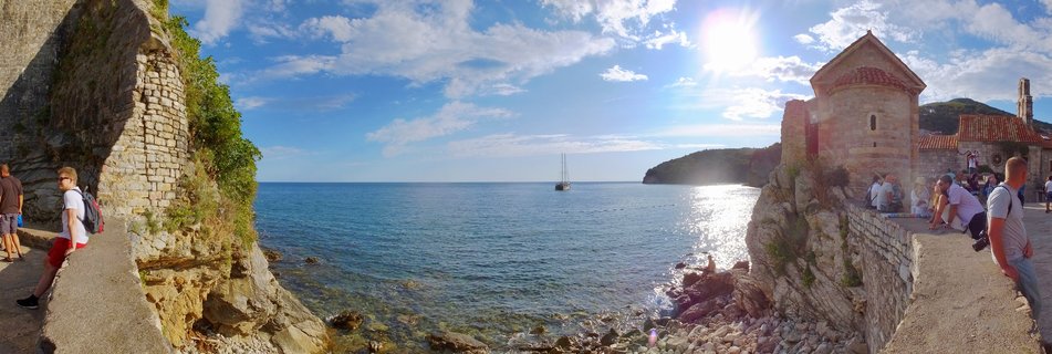 Panoramic view of Budva Old Town showing medieval stone walls, a red-tiled church, tourists walking on fortifications, rocky coastline, sailboat on the Adriatic Sea, and bright sun in blue sky with clouds