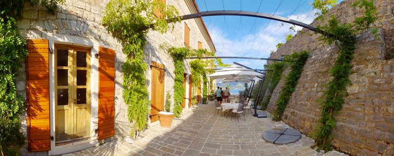 Outdoor stone courtyard terrace between historic buildings with wooden shutters, green climbing vines on walls, dining tables with white umbrellas, metal arch overhead, blue sky, people in background