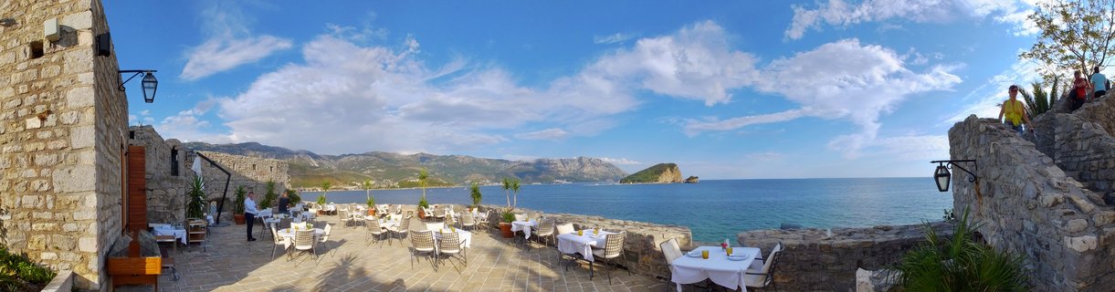 Outdoor restaurant terrace with white tablecloths and chairs on stone pavement, overlooking blue Adriatic Sea with island and mountains in distance under cloudy sky