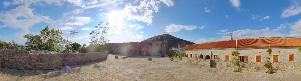 Wide panoramic view of a historic stone courtyard with terracotta-roofed buildings, stone walls, cobblestone pavement, and a flagpole with the Montenegrin flag, with a green hill and blue sky with clouds in the background