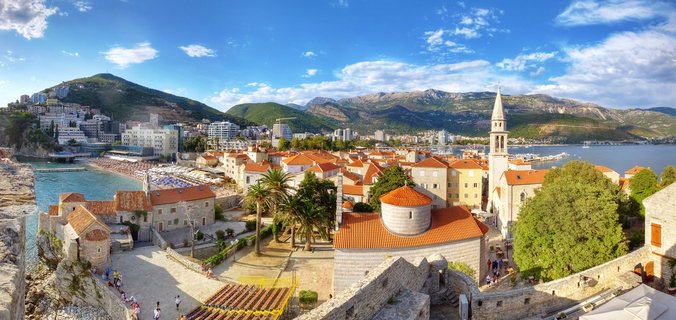 Panoramic aerial view showing Budva Old Town with orange-tiled roofs, a tall church bell tower, stone fortress walls, beach with umbrellas, blue sea, and green mountains under partly cloudy sky in Montenegro