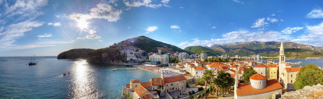 Aerial panoramic view of Budva Old Town showing red-tiled roofs, church bell tower, Adriatic Sea with boats, beach, and mountains under blue sky with sun