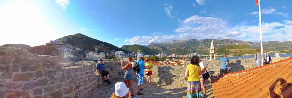 Tourists walking on stone fortress walls overlooking Budva Old Town with church bell tower, red-tiled roofs, mountains and Adriatic Sea in Montenegro