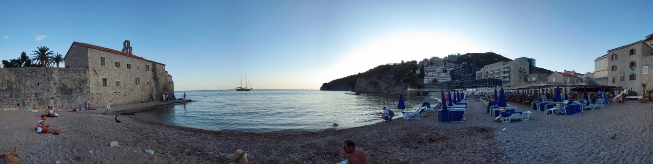 Panoramic beach scene showing historic stone buildings with bell tower on the left, calm blue sea with sailboat in center, and beach resort with blue umbrellas and lounge chairs on the right, under clear sky at sunset in Budva, Montenegro