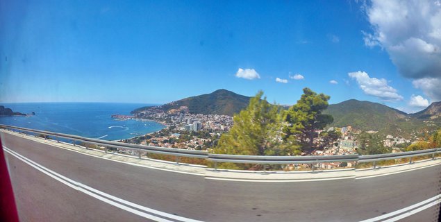 Panoramic coastal view showing a city built along hillsides next to blue sea, with a highway guardrail in foreground and mountains in the background under partly cloudy sky