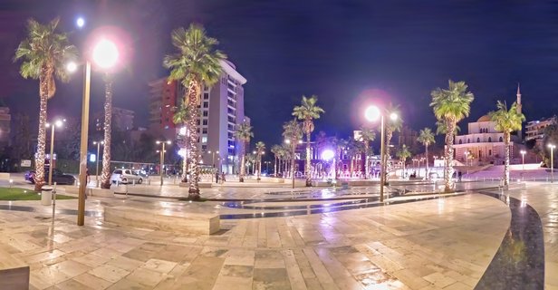 Night photograph of an illuminated urban plaza with palm trees, street lamps, marble paving, and a mosque with minaret in Durres, Albania