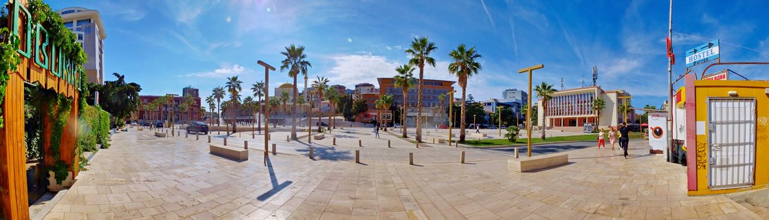 Wide panoramic view of a paved public plaza in Durres, Albania with palm trees, modern street lights, buildings in the background, and people walking under a blue sky