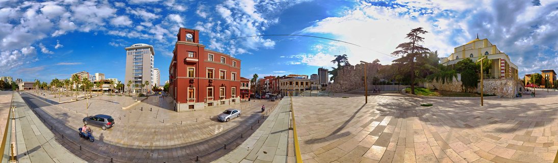 Panoramic street view of Durres city square with red clock tower building, ancient stone walls, yellow church, modern buildings, palm trees, cars and scooter on paved plaza under blue cloudy sky
