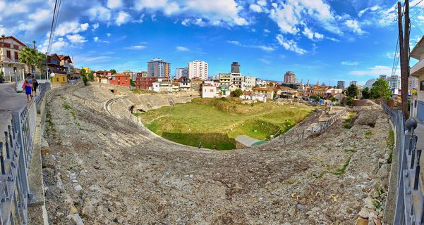 Panoramic view of the ancient Roman amphitheatre in Durrës, Albania, showing stone seating tiers surrounding a grassy central area with modern buildings and blue sky in the background