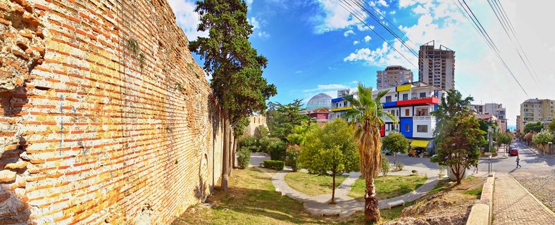 Ancient red brick Roman fortification walls on the left with modern colorful buildings, palm trees, and city streets visible on the right under blue sky in Durres, Albania