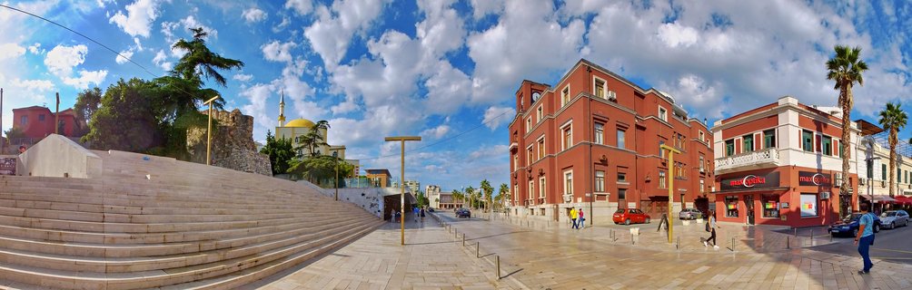 Panoramic street view of Durres city center with wide stone stairs, red buildings, palm trees, and a mosque with golden dome under blue sky with clouds