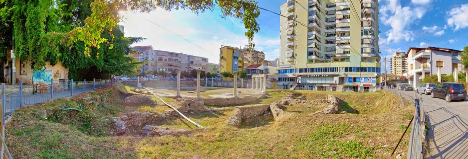 Archaeological excavation site showing ancient Roman stone columns and foundation walls with grass-covered ground, surrounded by modern multi-story apartment buildings under a blue sky with scattered clouds in Durrës, Albania.