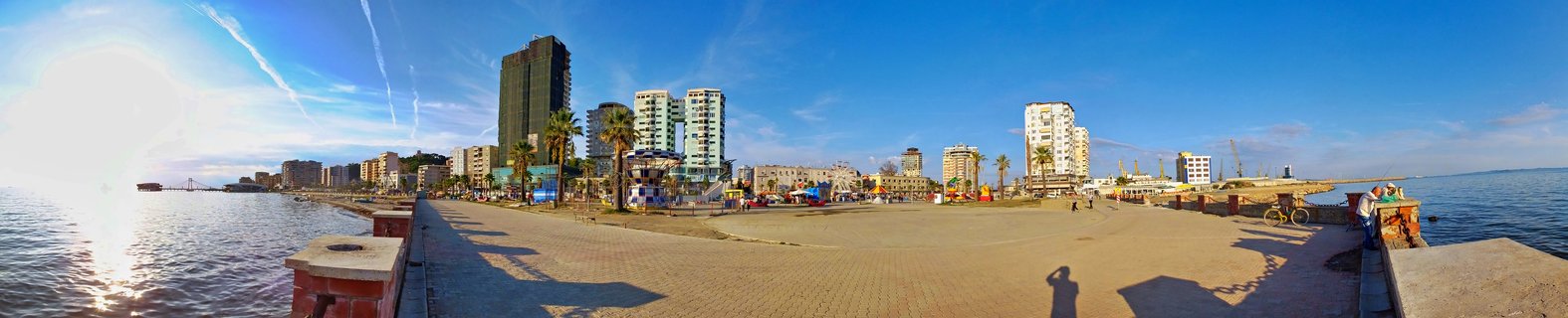 Panoramic waterfront scene showing Durres city skyline with tall buildings, palm trees, and paved promenade along the Adriatic Sea under blue sky with sun visible