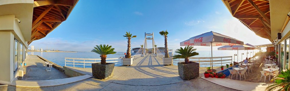 Panoramic waterfront restaurant terrace in Durres Albania with white cable-stayed pier, palm trees, outdoor dining tables with Coca-Cola umbrellas, and Adriatic Sea under clear blue sky