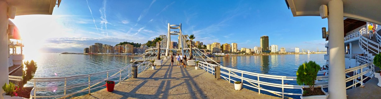 Wide panoramic view of a white cable-stayed pedestrian bridge at a marina with people walking, waterfront buildings along the coast, blue sky and calm water at sunset in Durres, Albania