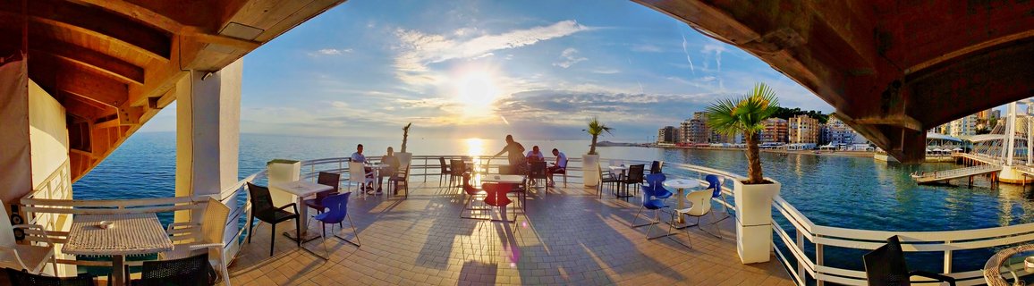 Outdoor restaurant terrace with people dining at tables, overlooking a harbor with buildings and the sea at sunset, framed by wooden beams overhead