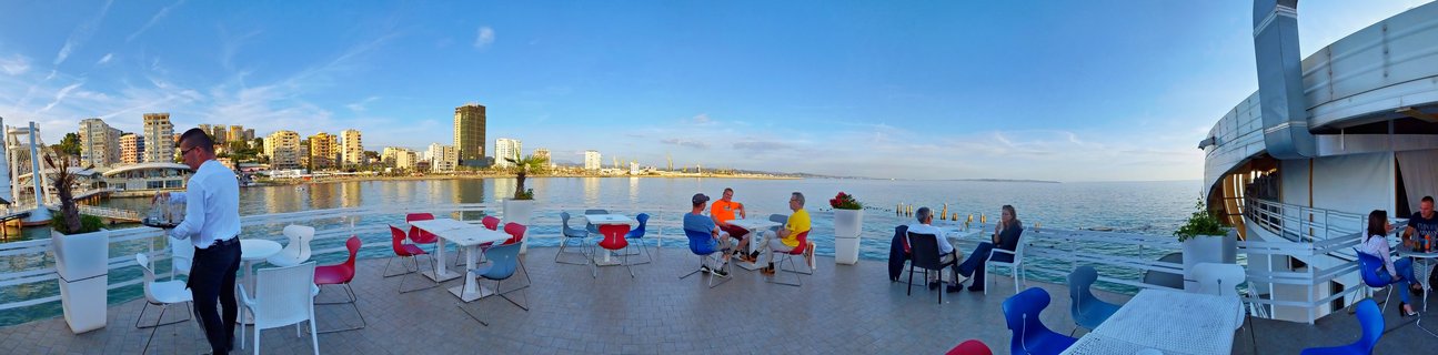 People sitting at colorful tables on a waterfront café terrace overlooking the Adriatic Sea in Durres, Albania, with city buildings visible across the bay under a blue sky