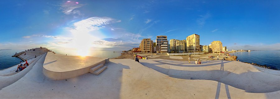 Panoramic sunset view of Durres waterfront with concrete promenade, people sitting on curved steps, modern buildings, and Adriatic Sea under blue sky with golden sunlight