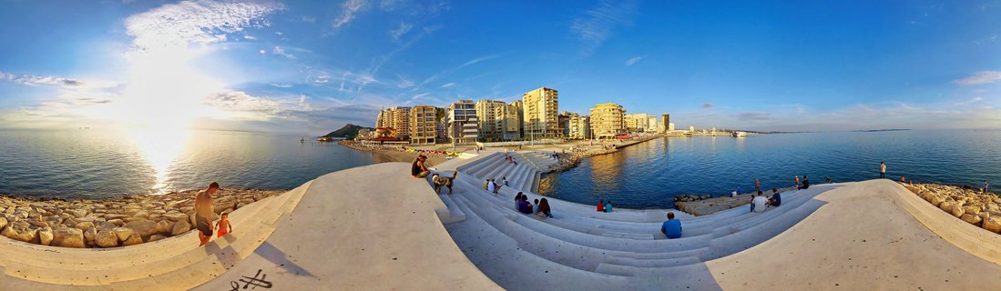 Wide panoramic view of people sitting on curved concrete amphitheater steps at Durrës waterfront during sunset, with apartment buildings along the coastline and the Adriatic Sea