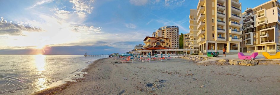 Panoramic beach scene at sunset in Durrës, Albania showing hotels, beach umbrellas, and colorful decorative bicycles along the Adriatic Sea shoreline