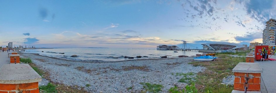 Wide panoramic photograph of Durrës beach and waterfront in Albania showing the Adriatic Sea, modern buildings on piers, pebble shoreline, promenade with pedestrians, and high-rise buildings under partly cloudy evening sky