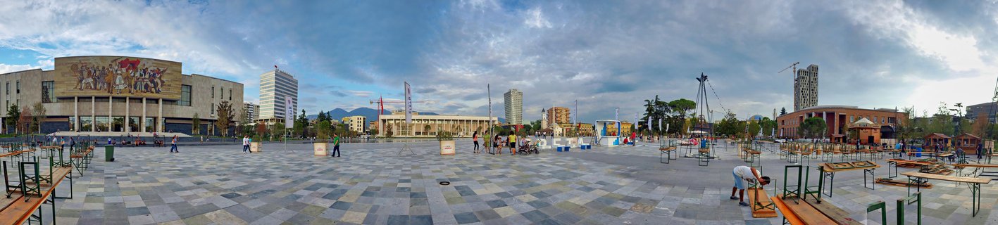 Wide panoramic view of Skanderbeg Square showing the National Historical Museum with large mosaic, paved plaza with people walking, outdoor seating, modern buildings and construction cranes under cloudy sky