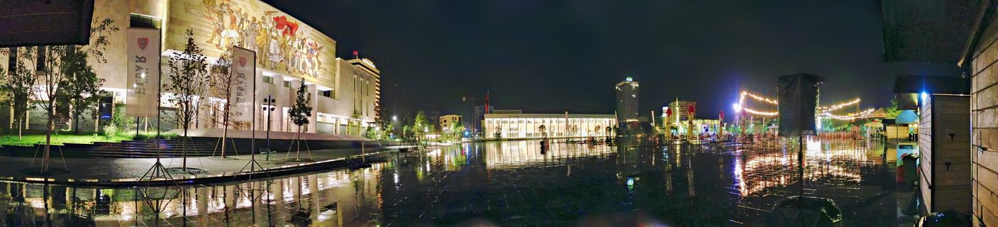 Night panoramic view of Skanderbeg Square in Tirana showing the National Historical Museum with illuminated mosaic facade, wet reflective pavement, string lights, and modern buildings in the background