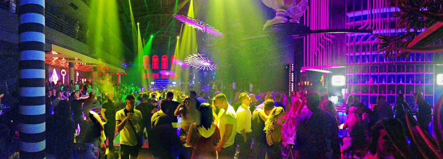 Crowded indoor nightclub with people dancing and socializing under green laser beams and purple neon lighting, featuring modern decorative elements and bar area