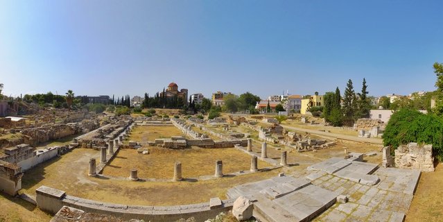 Panoramic view of ancient Greek archaeological ruins with stone column bases, surrounded by trees and modern Athens buildings, with a Byzantine church dome visible in the distance under clear blue sky