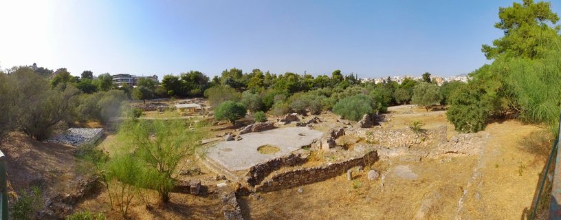 Panoramic view of ancient Greek archaeological ruins showing stone foundation walls, dry grass, olive trees, and Athens cityscape in the background under clear blue sky