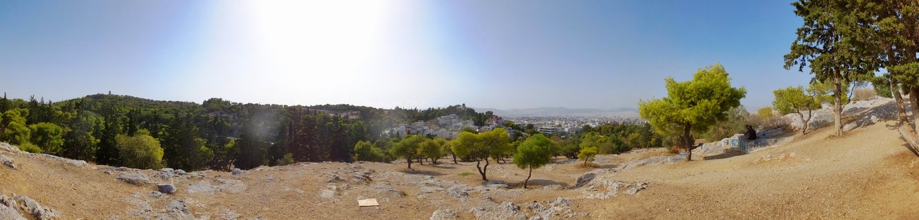 Wide panoramic photograph showing rocky archaeological terrain with scattered stones in foreground, green pine trees in midground, and Athens cityscape extending to distant mountains under bright blue sky