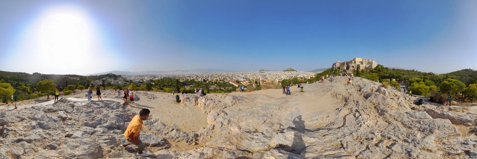 Wide panoramic view from Pnyx Hill showing tourists on rocky terrain with Athens cityscape and the Acropolis visible in the distance under bright blue sky