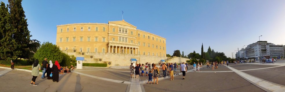 Wide view of the yellow Hellenic Parliament building with Greek flag, columns, and tourists walking across paved Syntagma Square under clear blue sky