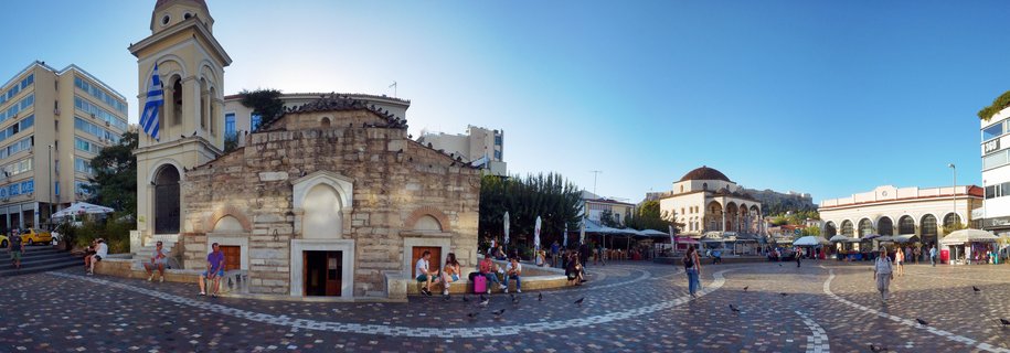 A panoramic view of Monastiraki Square in Athens showing the stone Church of Pantanassa with a Greek flag on its bell tower, people walking and sitting in the cobblestone plaza, market stalls with umbrellas, and the Acropolis hill in the background under a clear blue sky