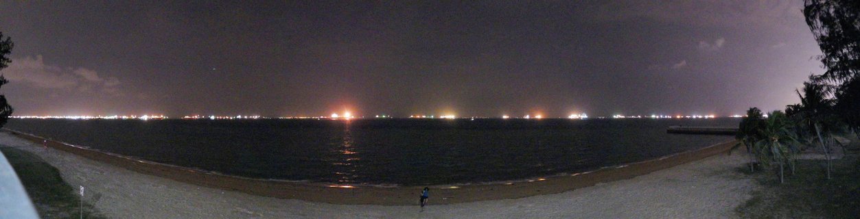 Night panoramic photograph showing a dark beach with palm trees, calm water reflecting distant ship lights along the horizon under a dark sky with one person visible on the sand.