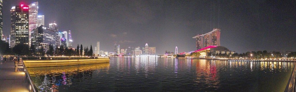 Nighttime panoramic view of Singapore's Marina Bay showing illuminated skyscrapers, Marina Bay Sands hotel, and city lights reflecting on water with waterfront walkway in foreground